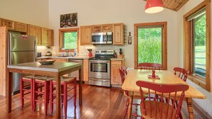 A kitchen with wooden cabinets, a stainless steel refrigerator, a microwave, an oven, and a dining area with a table and chairs. - Songbird Cabin | Hocking Hills, Private Trails (South Bloomingville)
