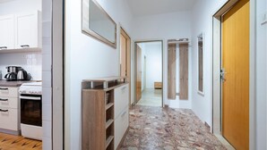 Un couloir étroit, avec une cuisine d’un côté, comprenant un fourneau, une machine à café et des placards. On y trouve des portes en bois et un sol carrelé.
