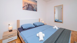 Une chambre d’hôtel moderne avec un lit en bois, une table de chevet, une lampe de chevet, un grand miroir et un tableau représentant un coucher de soleil accroché au mur.