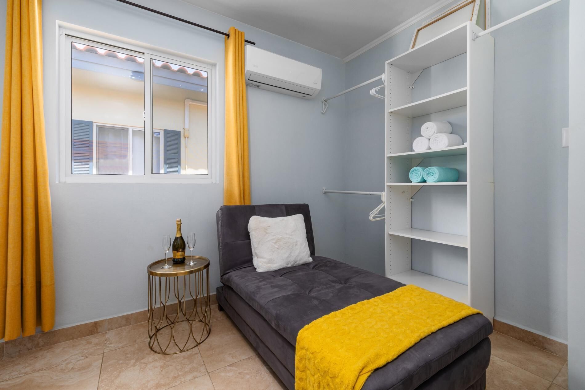 Une chambre à coucher de taille modeste, comprenant un lit, une petite table ronde avec une bouteille et des verres, une armoire avec des étagères et une fenêtre avec des rideaux.