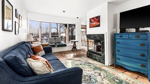 Una sala moderna con un sofá azul, una chimenea, un televisor y un ventanal con vistas a árboles.