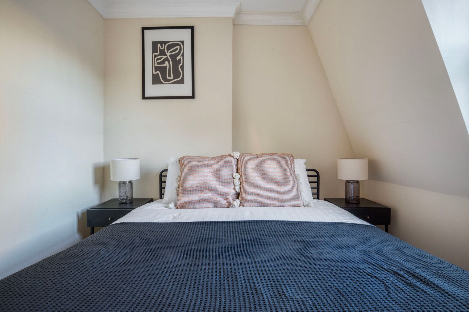 Ein Schlafzimmer mit einem Bett, zwei Nachttischen, zwei Lampen, einem gerahmten Kunstwerk an der Wand und einer schrägen Decke.