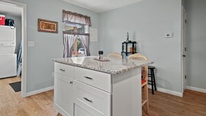 A compact kitchen with a granite countertop, white cabinetry, and a window with curtains. - Escape at Deep Hole Hideaway | Chic 2BR Retreat w/ Full Kitchen, Daybed + Sleeper Sofa, Pet Friendly (Chincoteague Island)