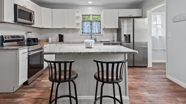 A modern kitchen with white cabinets, a granite countertop, stainless steel appliances, and two bar stools. - Serenity at Deep Hole Hideaway | Cozy Retreat w/ Deck, Firepit, Full Kitchen & Pet Friendly (Chincoteague Island)