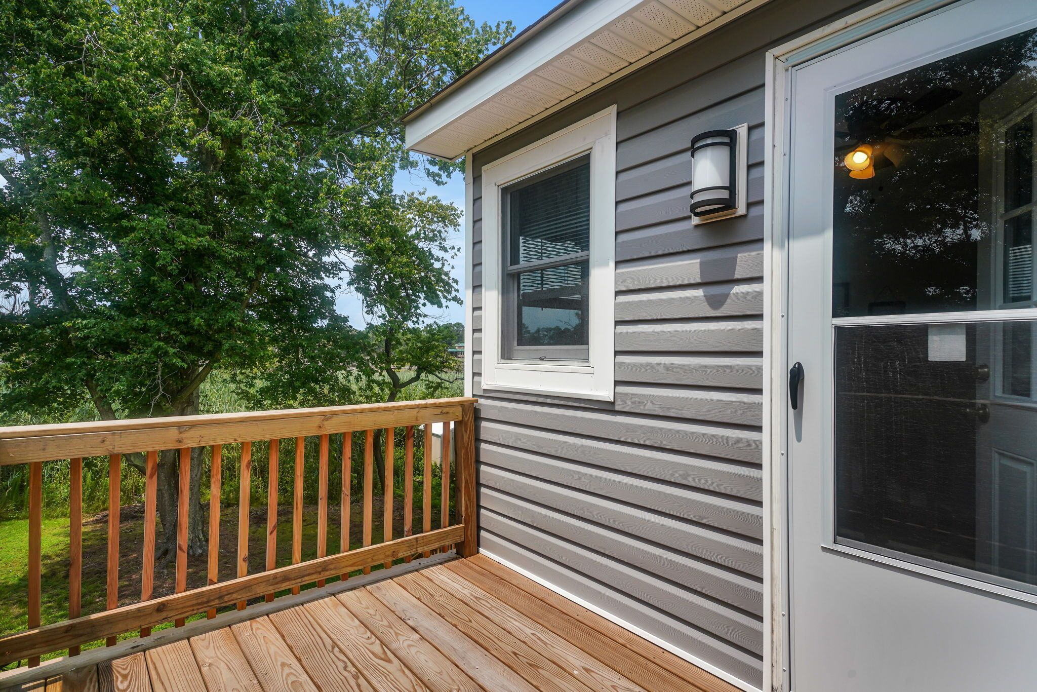 A wooden deck with a metal railing, a gray building with a window and a door, and a tree in the background.