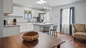 A modern kitchen with white cabinets, a central island, and a dining area with a wooden table and chairs. - Serenity at Deep Hole Hideaway | Cozy Retreat w/ Deck, Firepit, Full Kitchen & Pet Friendly (Chincoteague Island)