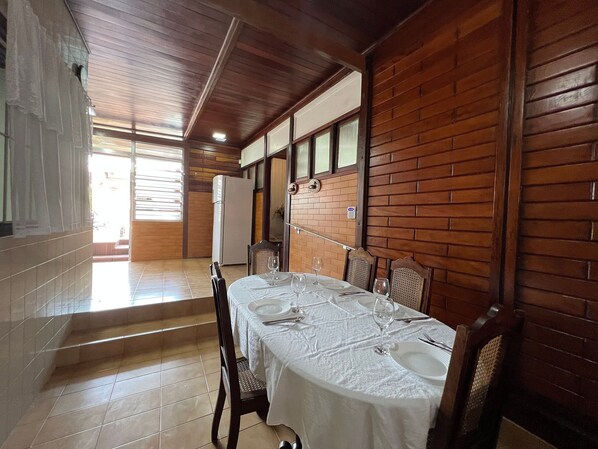 A dining area with a table set for four, wooden walls, a refrigerator, and a window with blinds. - 150m Praia -Ar condicionado -Petfriendly (Peruíbe)
