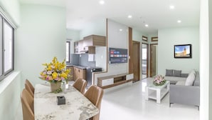 A modern living room with a grey sofa, a dining table with chairs, and a kitchen area with a TV. - Happy Hotel Apartment (Cam Ly - Da Lat)
