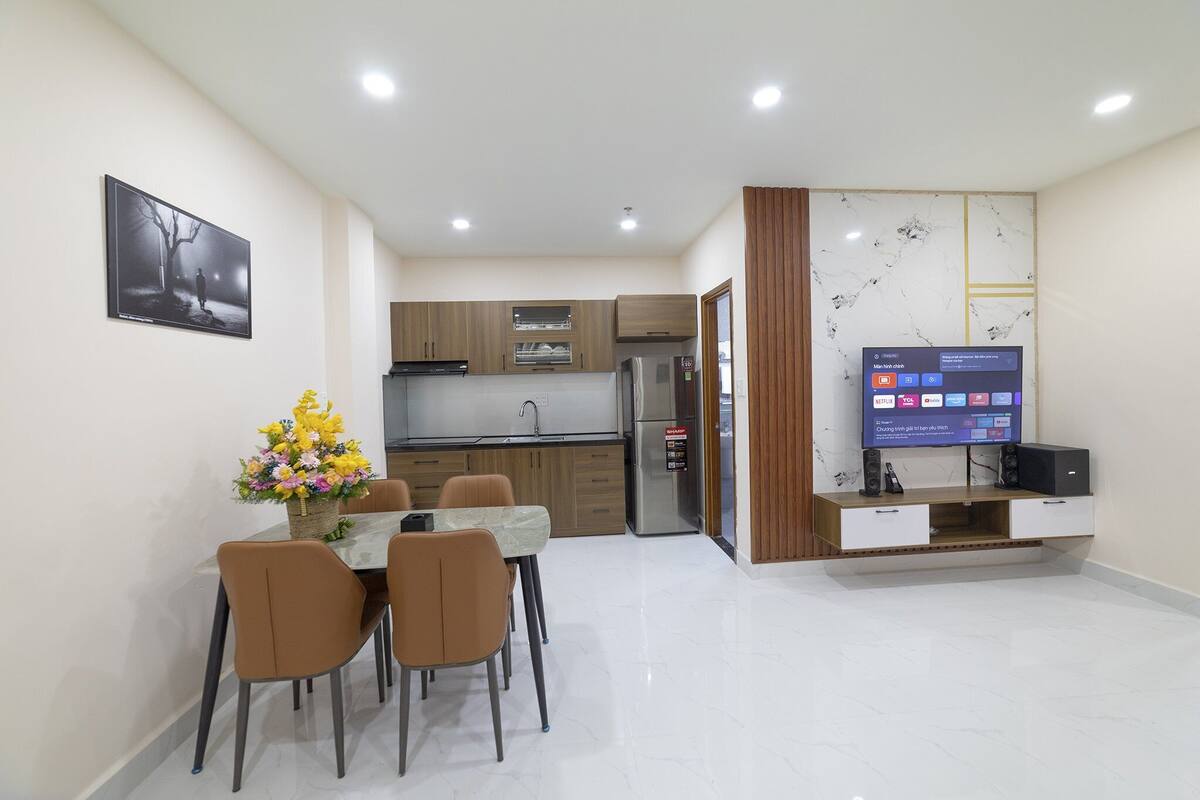 A modern kitchen with wooden cabinets, a built-in oven, and a refrigerator. There is a dining table with chairs and a bouquet of flowers. A flat-screen TV is mounted on the wall.