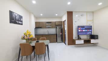 A modern kitchen with wooden cabinets, a built-in oven, and a refrigerator. There is a dining table with chairs and a bouquet of flowers. A flat-screen TV is mounted on the wall.