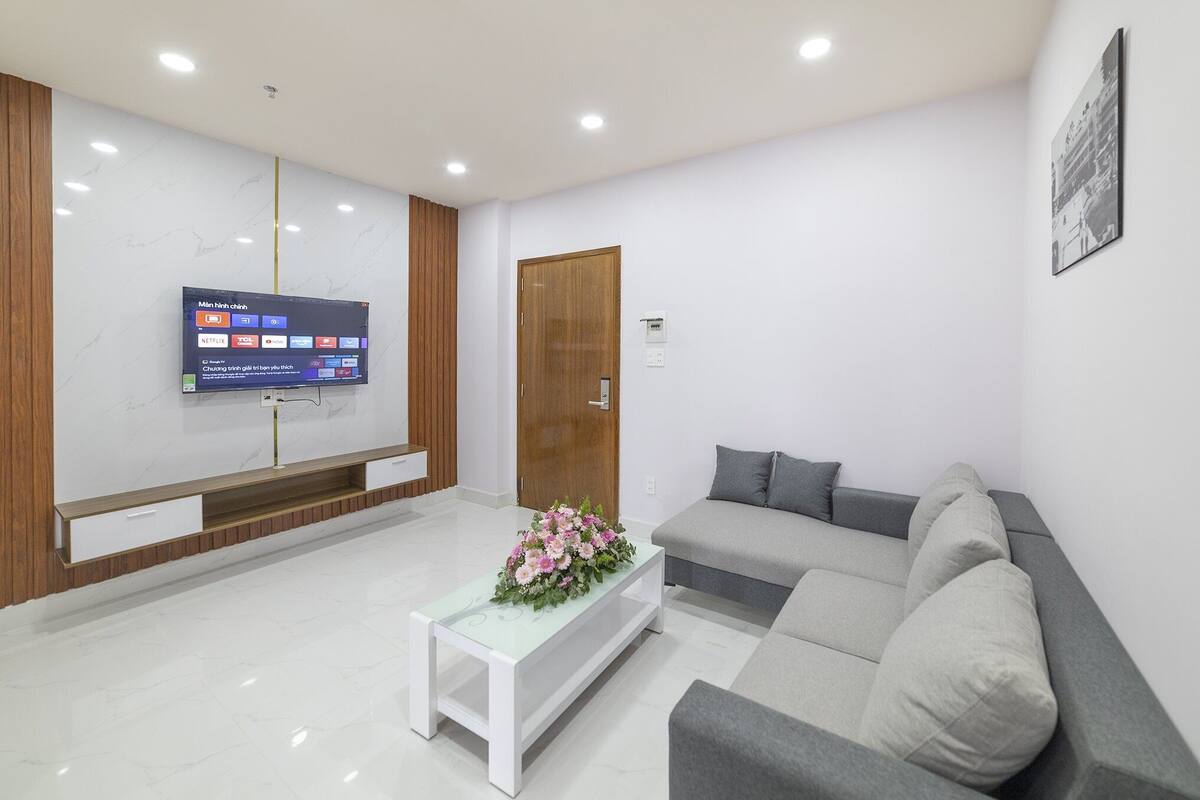 A modern living room with a flat-screen TV mounted on the wall, a grey sofa, a white coffee table with a floral centerpiece, and a wooden door.