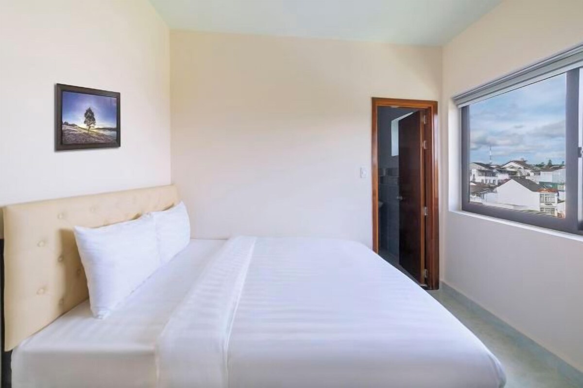 A hotel room with a bed, a window with a city view, and a door leading to another room.