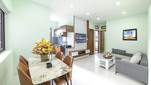 A modern living room with a dining area, a grey sofa, a TV mounted on the wall, and a kitchen in the background. - Happy Hotel Apartment (Cam Ly - Da Lat)