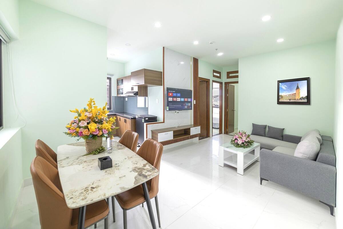 A modern living room with a dining area, a grey sofa, a TV mounted on the wall, and a kitchen in the background.