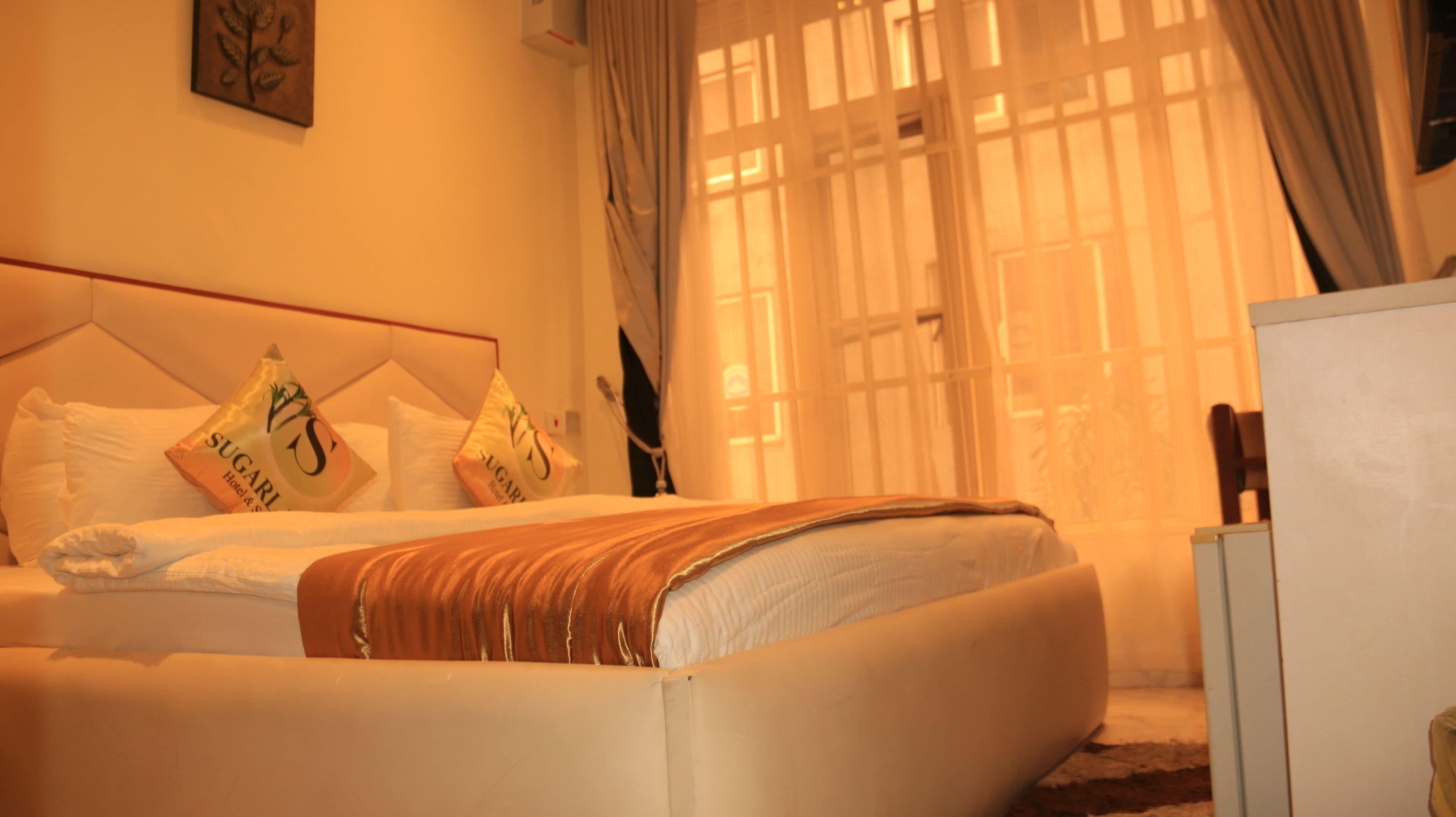 Una habitación de hotel con cama, almohadas, una colcha, una mesita de noche y un ventanal con cortinas.