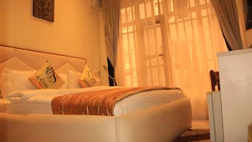 Una habitación de hotel con cama, almohadas, una colcha, una mesita de noche y un ventanal con cortinas.