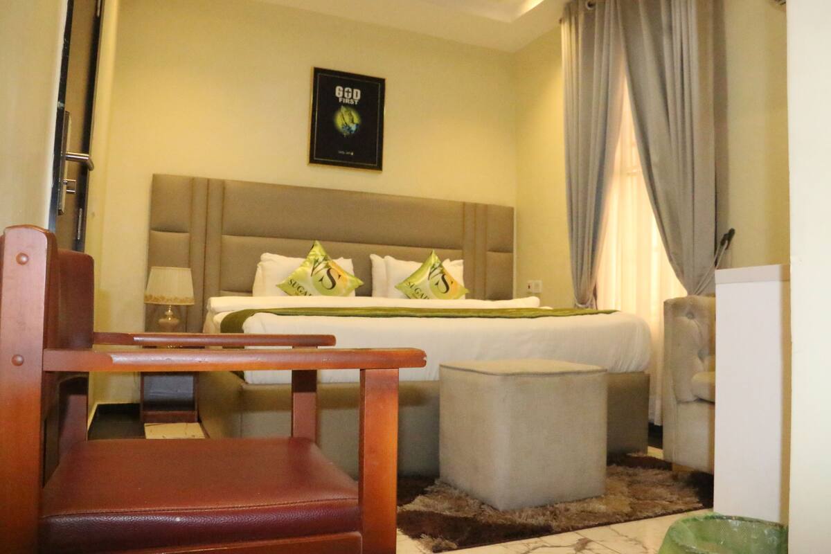 A hotel room with a bed, a wooden chair, a small table, a nightstand, and a lamp.