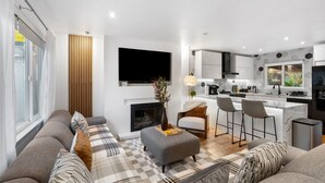 Een moderne woonkamer met een bank, een voetenbankje en een open haard. Er is een keuken met een bar en zitplaatsen.