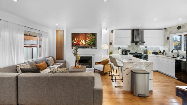 Een moderne woonkamer met een grijze bank, een eethoek met een witte tafel, en een keuken met witte kasten en een zwarte afzuigkap.