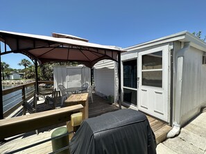 A covered outdoor patio with a table and chairs, a covered grill, and a white mobile home with a sliding door. - Cozy little Lake Front Park Model (Port Richey)
