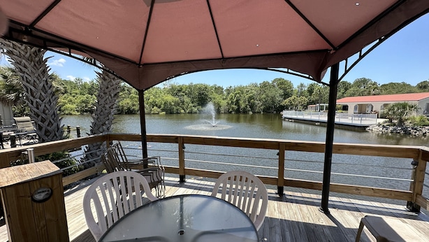 Terraza de madera con mesa y sillas, con vistas a un lago que tiene una fuente y un muelle para embarcaciones.