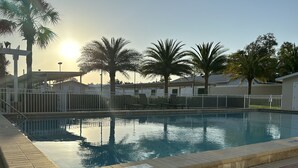 Una piscina con plataforma de azulejos, rodeada de palmeras y una valla blanca.