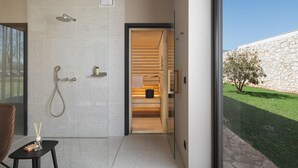 A modern bathroom with a shower, a small table with toiletries, and a view of a stone wall and garden outside. - Stunning home in Vodnjan with sauna (Vodnjan)