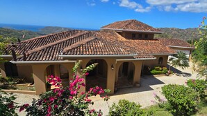 A Mediterranean-style villa with terracotta tiled roofs, arched doorways, and a courtyard with flowering plants. - Housekeeping•BBQ•Pool•4 TVs•Garden•Oceanview villa: Casa Sol Azul (Potrero)