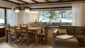 Un salon chaleureux avec une table à manger en bois, des chaises et un canapé. Une grande fenêtre offre une vue sur les arbres et les bâtiments.