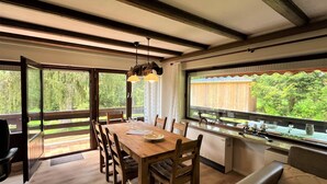 Un coin repas avec une table et des chaises en bois, un four encastré et une vue sur la verdure grâce à de grandes fenêtres.
