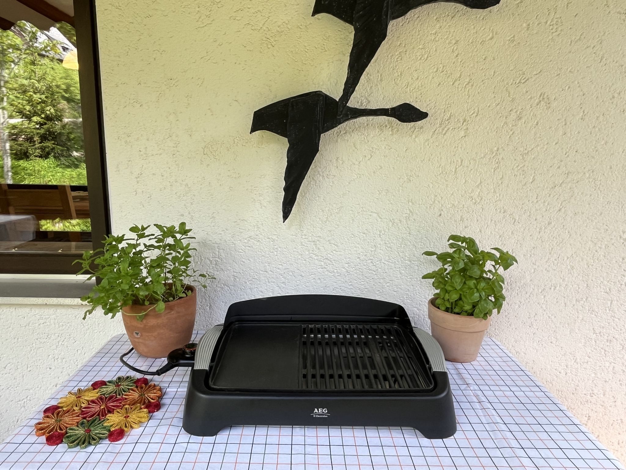 A grill on a tiled patio with two potted plants and a decorative flower arrangement.