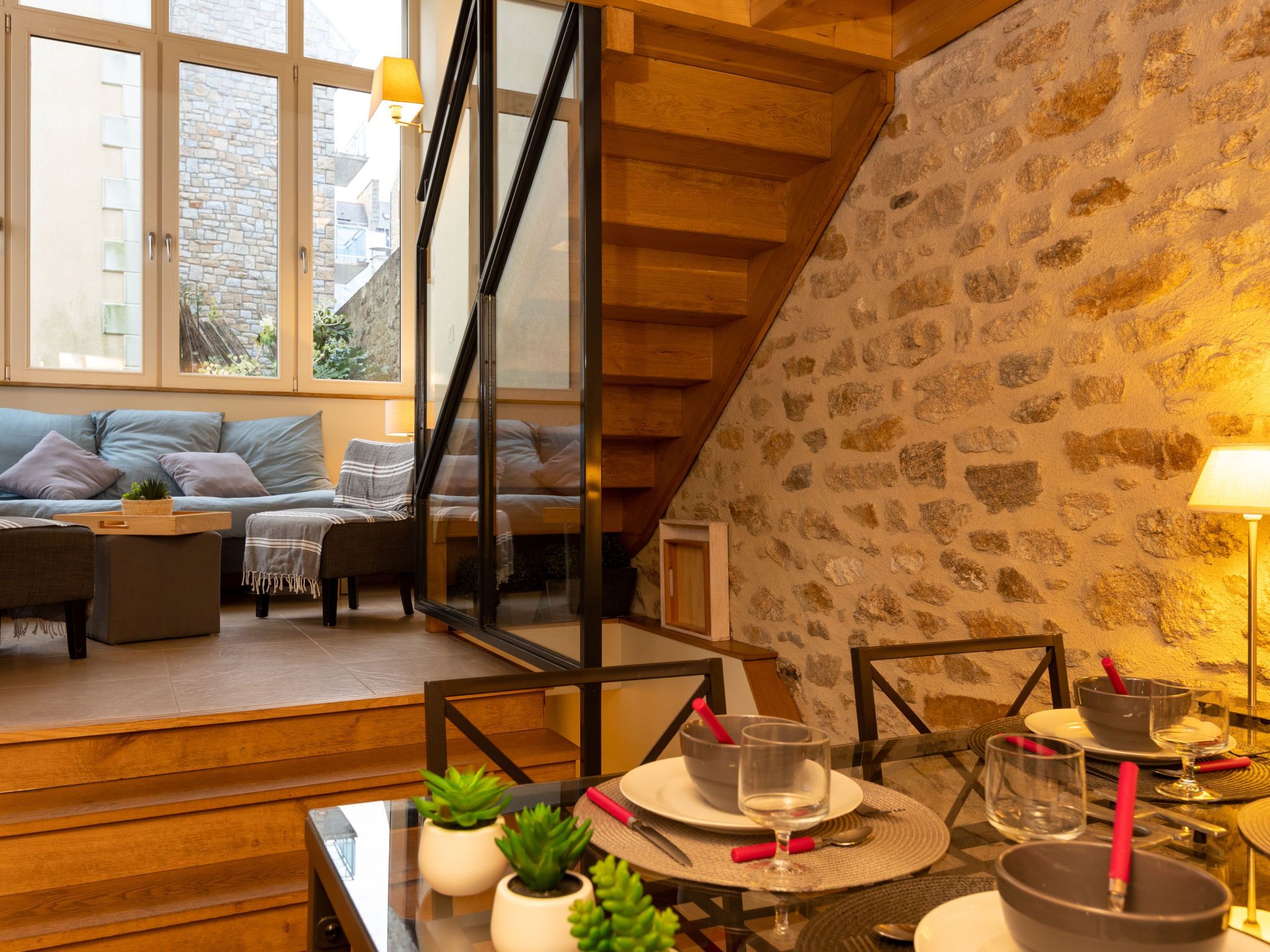 A cozy living room with a stone wall, wooden staircase, and a dining area with a glass table.