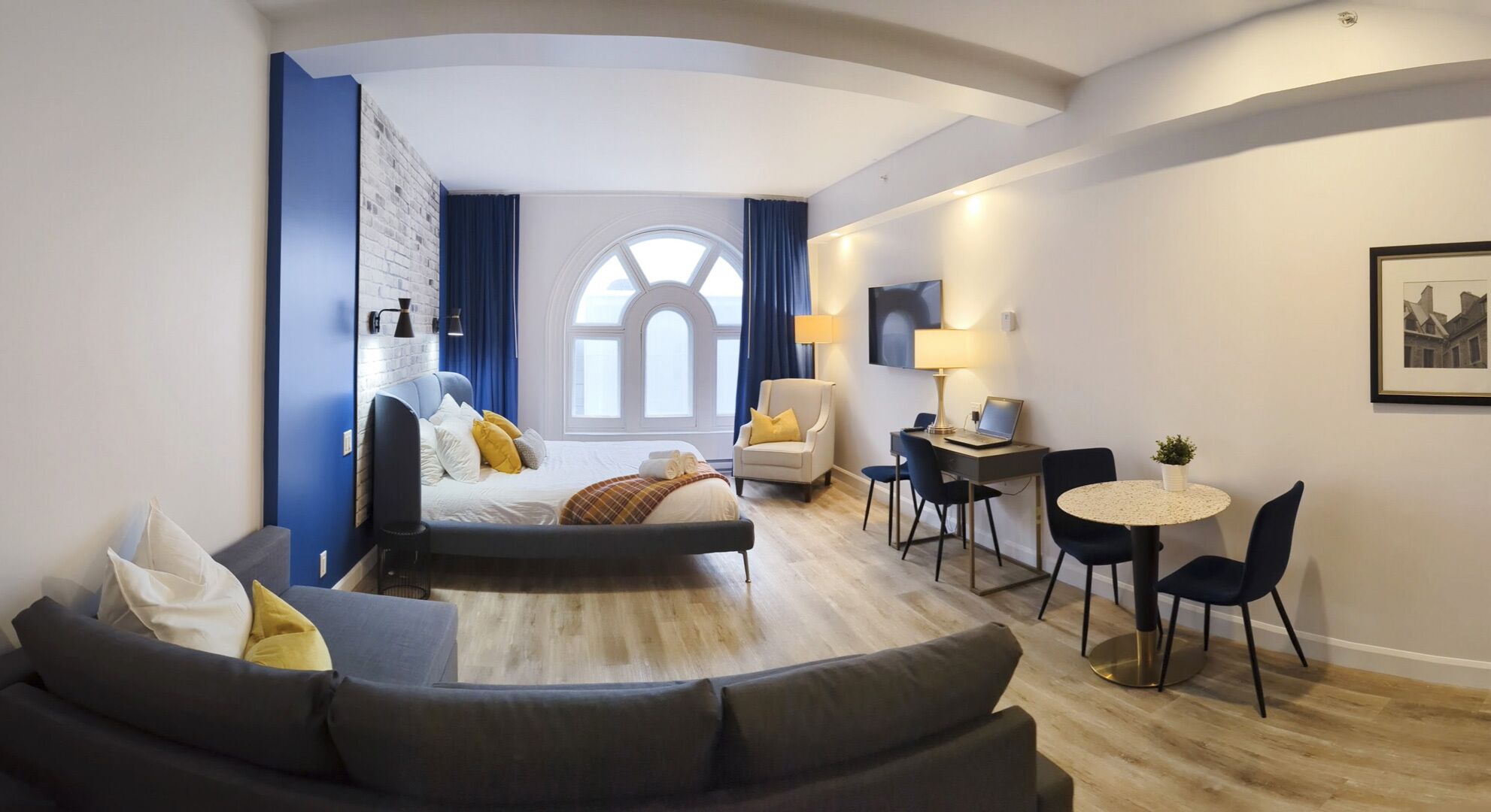 A modern hotel room with a curved sofa, a small round table, a desk with a laptop, and a large window with curtains.