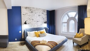 A modern bedroom with a large bed, a brick accent wall, a view of the city through a large window, and a small armchair. - Modern Loft - Downtown Québec City (Québec)