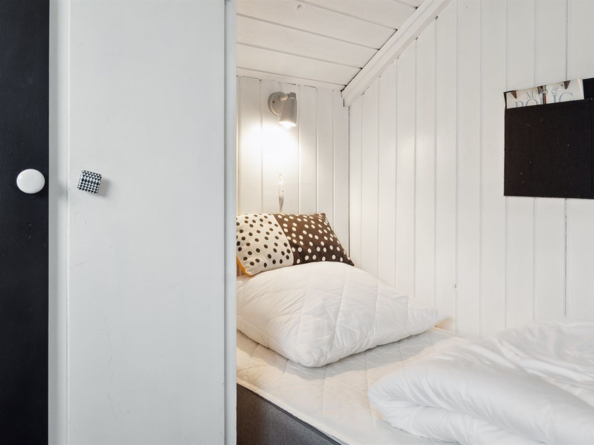 A small, single-bed room with white walls and a wooden ceiling. There is a bed with white and polka-dotted pillows, and a small, round, white light fixture above it.