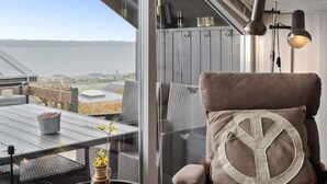 A cozy seating area with a view of a body of water, a table with a vase of flowers, and a lamp on the wall.