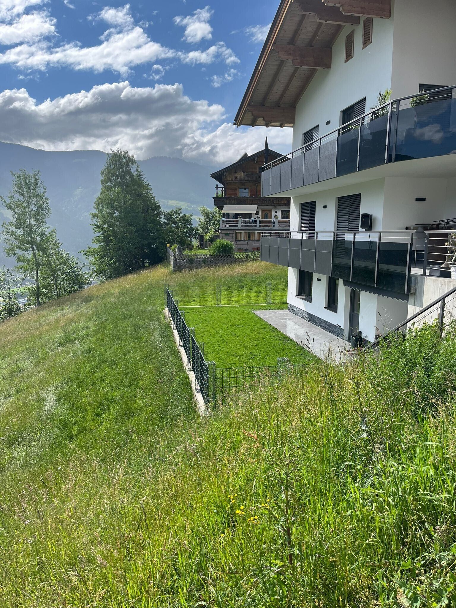 Ein modernes, zweistöckiges Haus mit Balkon, einer sanften Rasenfläche und Blick auf die Berge.