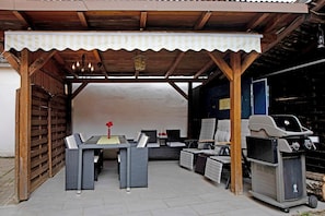 Een terras buiten met een overdekte zithoek, een barbecue en een tafel met stoelen.