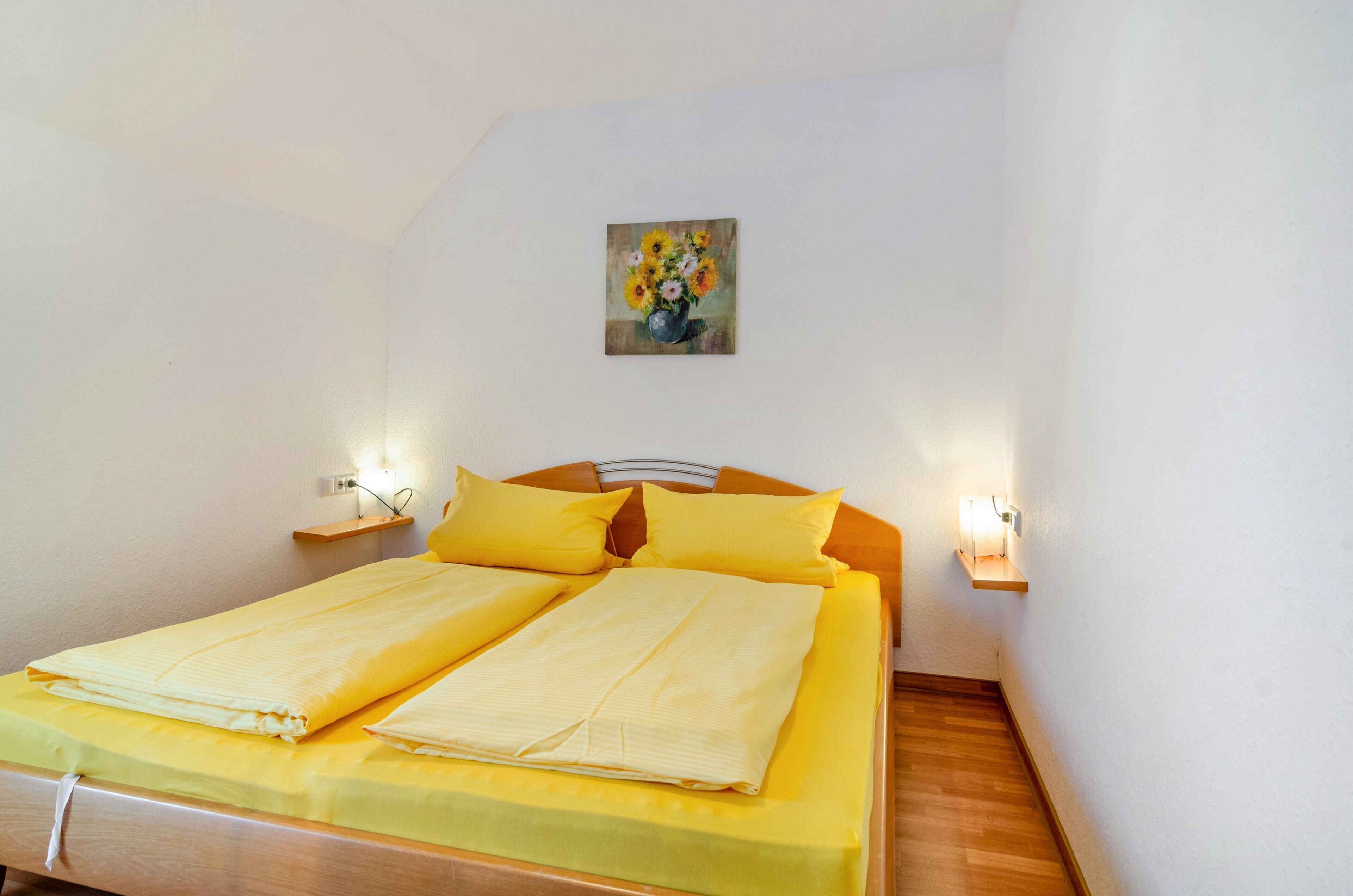 Ein Einzelbett mit gelber Bettwäsche, ein Bild an der Wand und Nachttischlampen.