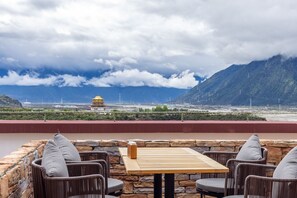 Une terrasse dotée de chaises en osier et d’une table en bois, offrant une vue sur un paysage montagneux et sur les contours d’une ville lointaine.