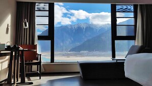 Hotelkamer met een groot raam met uitzicht op de bergen, een bed, een bureau met stoel en een kleine tafel met waterkoker.