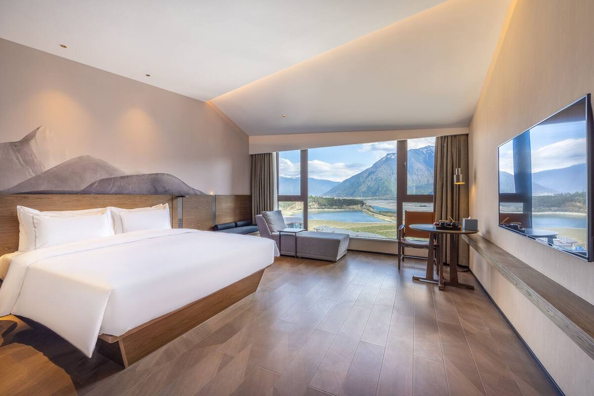A modern hotel room with a large bed, a seating area, and a view of mountains and a lake.