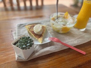 A slice of cake with a scoop of ice cream on a white plate, a glass of orange juice, and a small potted succulent plant on a wooden table.