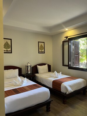 A hotel room with two single beds, each with a wooden headboard, white bedding, and a brown throw blanket. There are framed pictures of trees on the walls, and a window with wooden shutters.