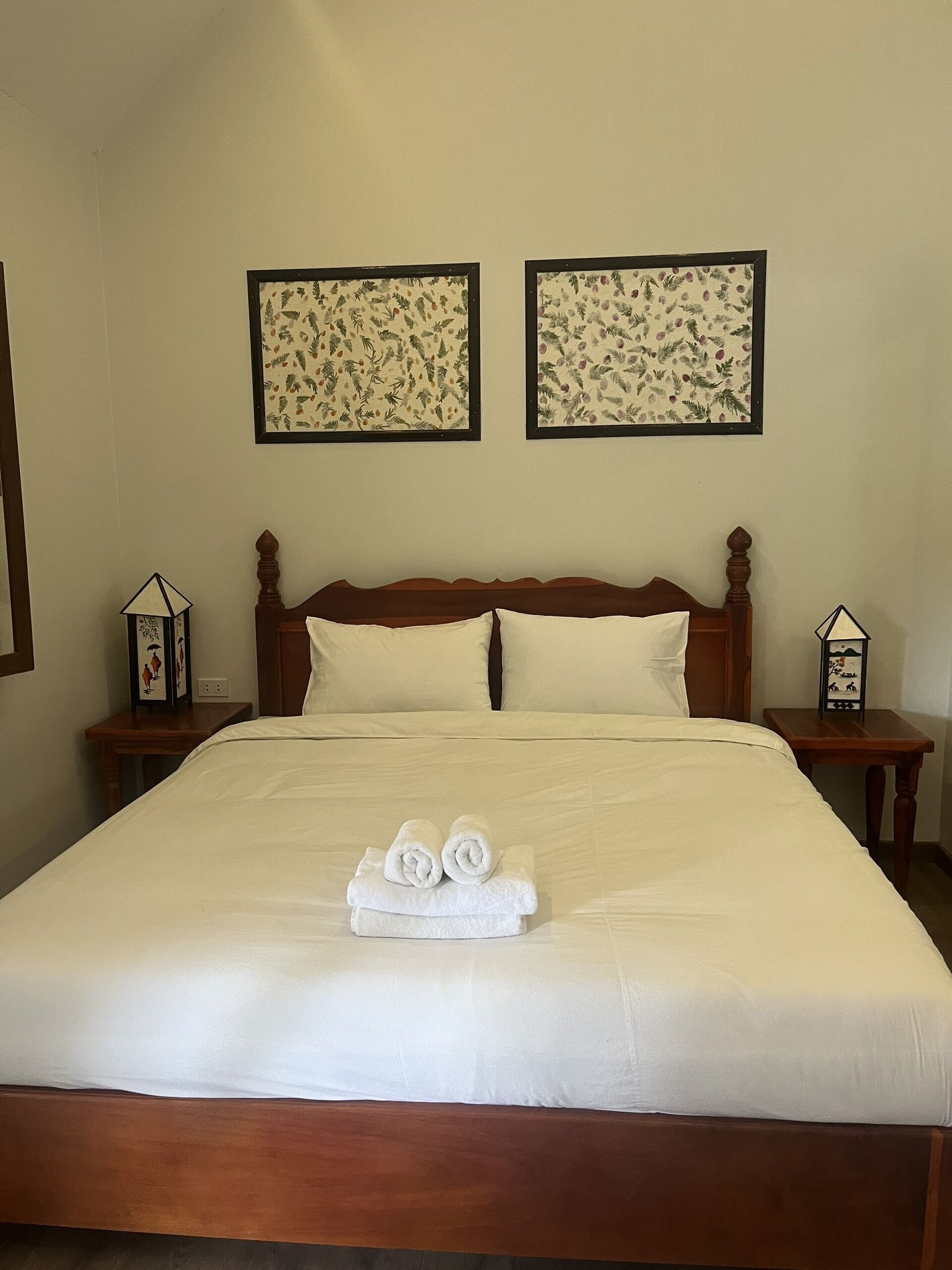 Un lit bien fait, avec du linge de lit blanc et des serviettes pliées, deux tableaux floraux encadrés accrochés au mur, et deux tables de chevet ornées de lanternes décoratives.