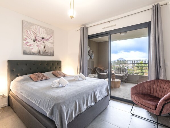 A modern bedroom with a large bed, a balcony with wicker chairs, and a city view.
