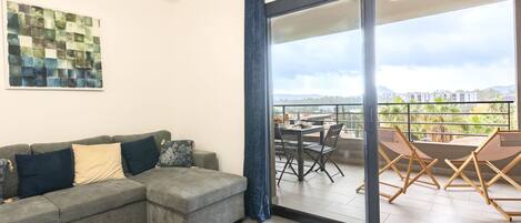 A modern living room with a grey sectional sofa, a wooden coffee table, and a dining area with a table and chairs. A large window offers a view of the cityscape and mountains.