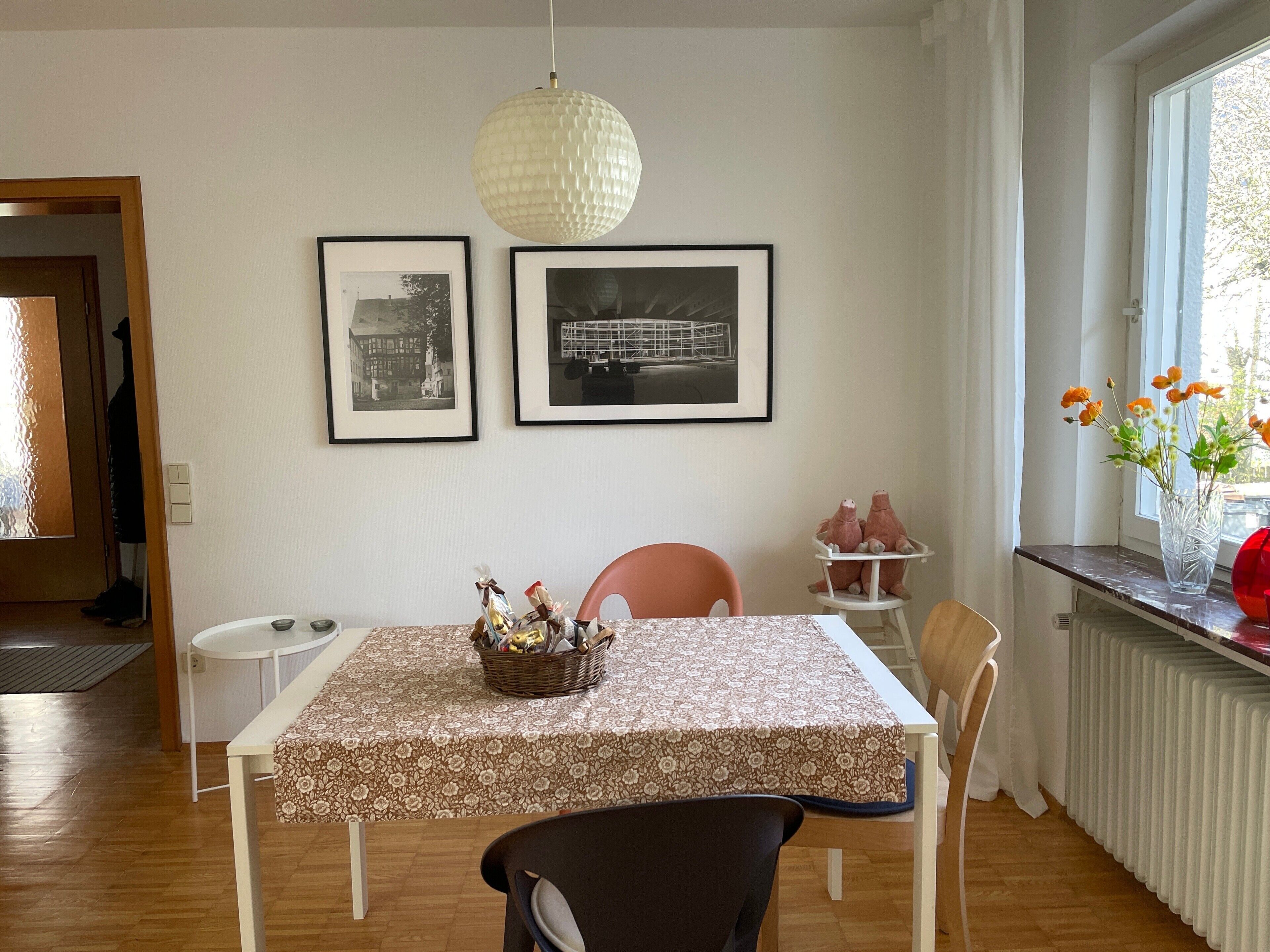 En matplats med ett bord, stolar, en hängande lampa, inramade tavlor och ett fönster med blommor.