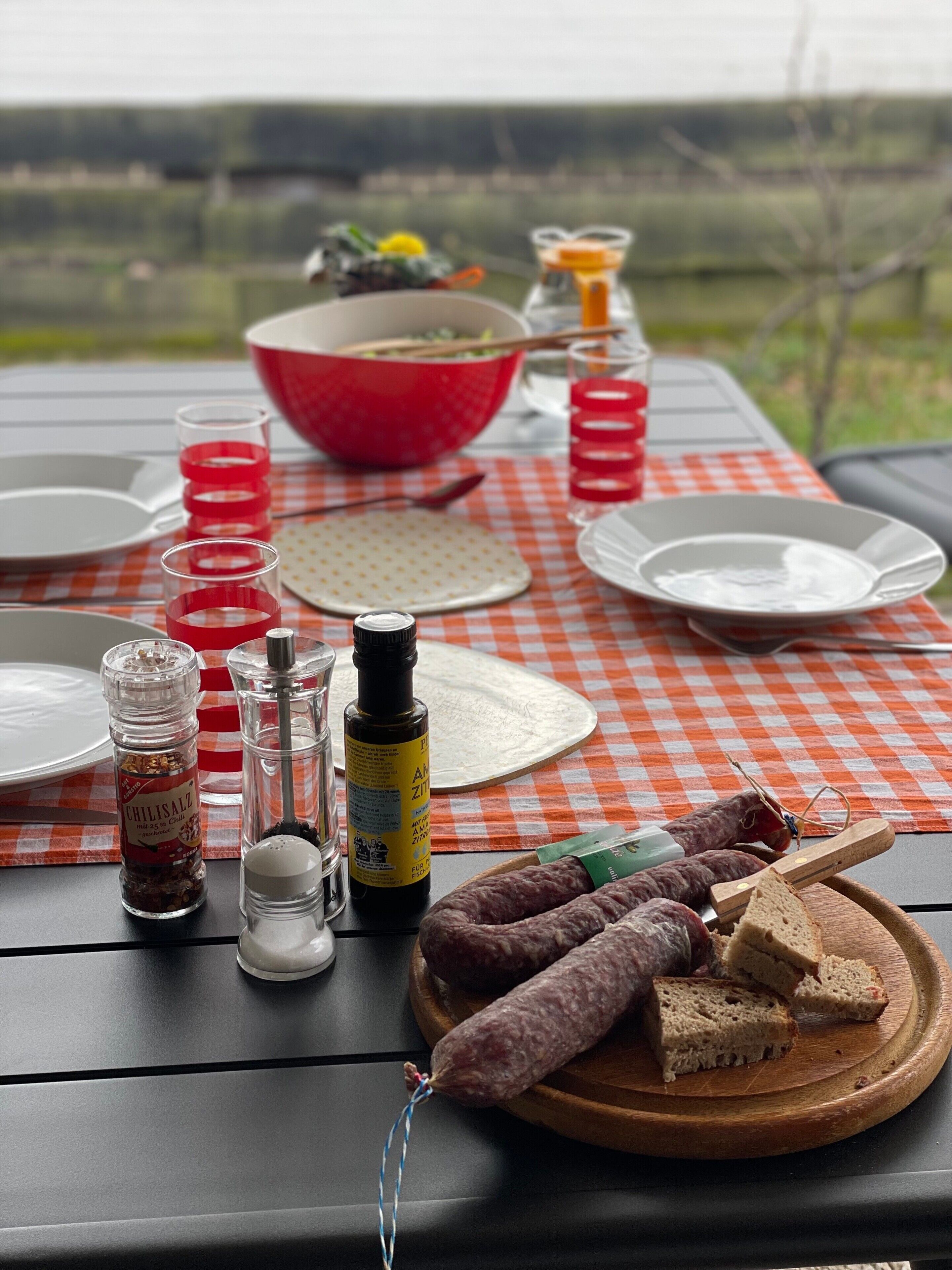 Una preparación para picnic con mantel a cuadros, platos, vasos, salsas y salchichas sobre una mesa.