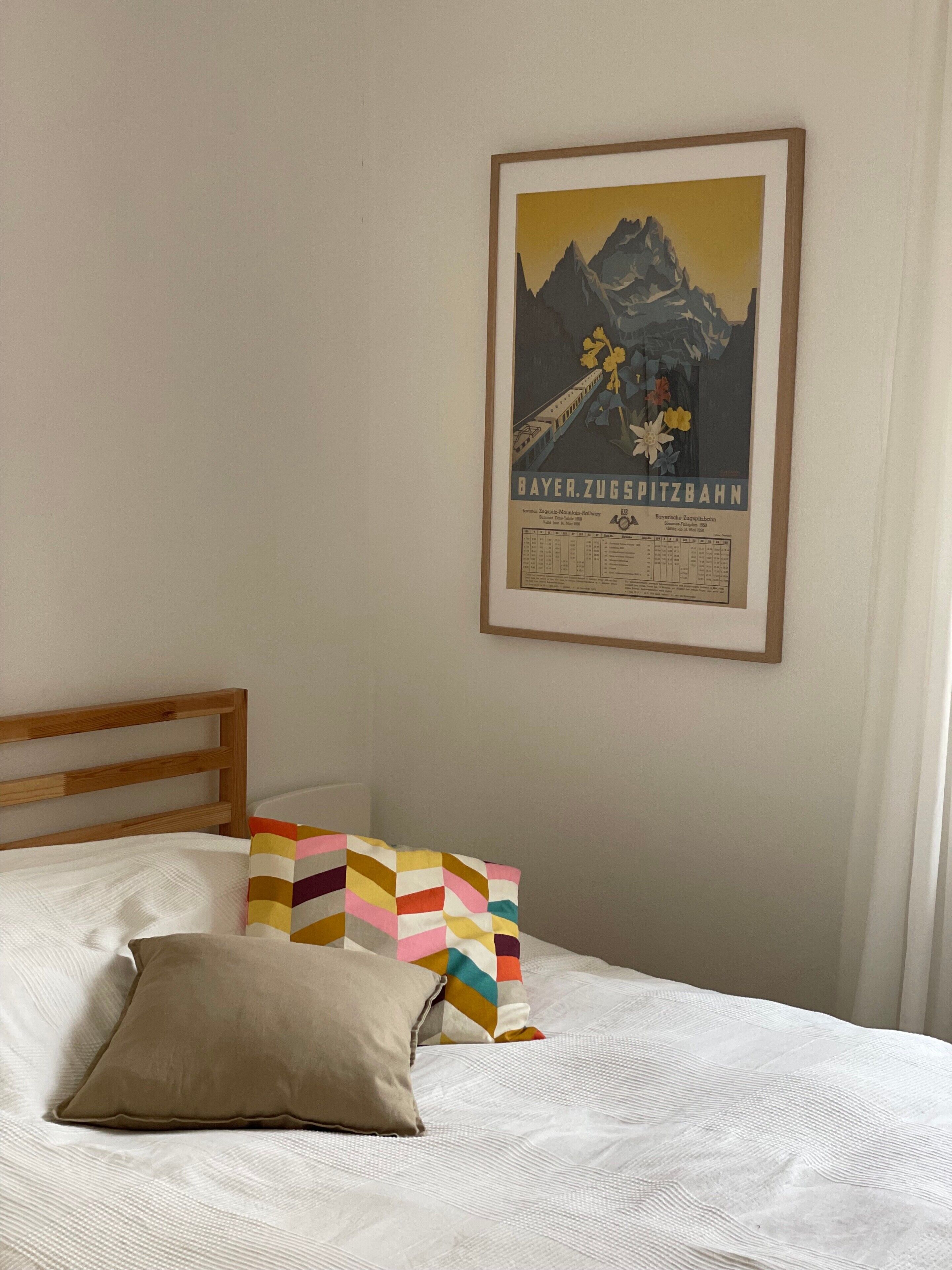 A bed with white bedding, a wooden headboard, and colorful pillows. A framed poster of a mountain landscape is on the wall.